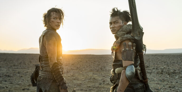 Two figures, a man and woman, wearing leather armor and bearing weapons, look towards the foreground as a sun behind them sets in a barren, desert landscape.