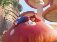 Screenshot from Illumination's The Super Mario Bros. Movie (2023). A 3D computer generated visual of a small man wearing a red outfit with a pair of blue, denim overalls cartoonishly smooshed against the cap of a large mushroom. A series of other large mushrooms and a rocky formation tower against the background.